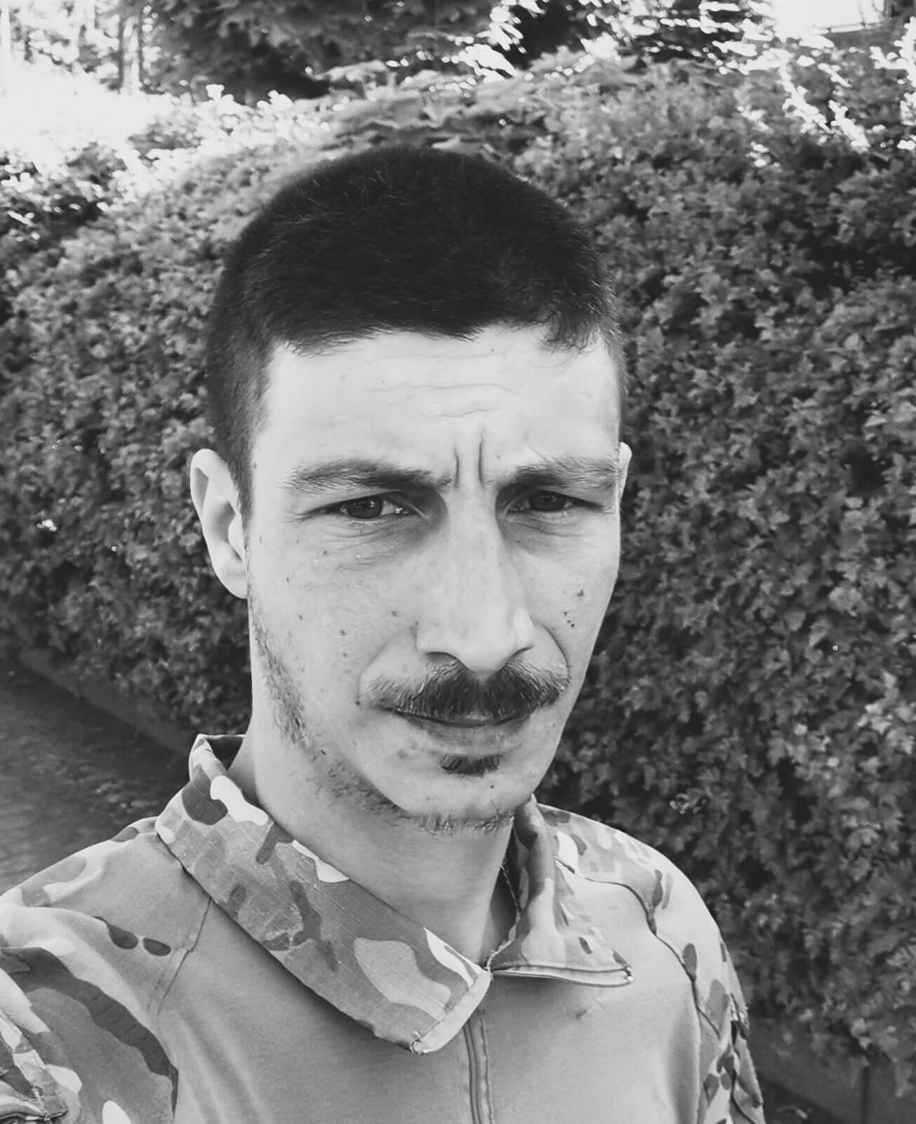 A man with a mustache and short hair wearing a camouflage jacket, standing outdoors in front of greenery.